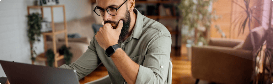 Homme barbu portant des lunettes, concentré en travaillant sur un ordinateur portable dans un environnement intérieur confortable.